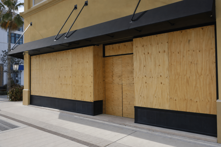 A storefront is covered with large plywood boards over the windows and doors. The building has a beige exterior with black awnings. No people are present.