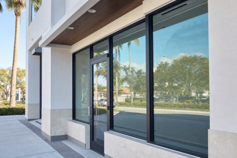 Glass storefront with a glass door and large windows reflecting trees, sidewalk, and buildings on a sunny day.