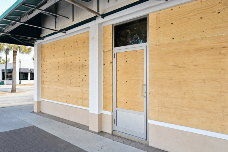 A storefront with windows and door covered by plywood boards, likely for protection. The sidewalk in front is empty and palm trees are visible in the background.