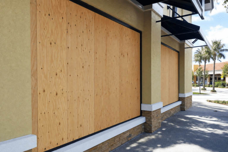 Storefront windows are covered with large plywood boards, possibly for protection or due to closure. The building is in a quiet outdoor shopping area with palm trees and a clear sky.