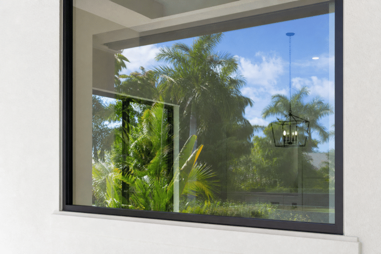 Large window reflecting palm trees, blue sky, and a hanging light fixture from inside the house.