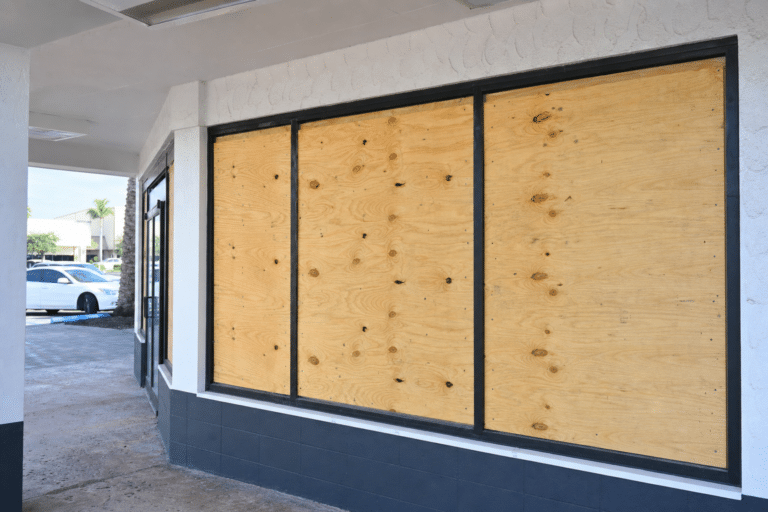 Storefront windows covered with sheets of plywood, possibly for protection or renovation. A white car is parked outside, and the area appears to be part of a shopping plaza.