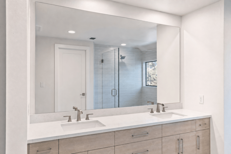Modern bathroom with a large mirror, double sinks with silver faucets, light wood cabinets, and a glass shower enclosure in the background.