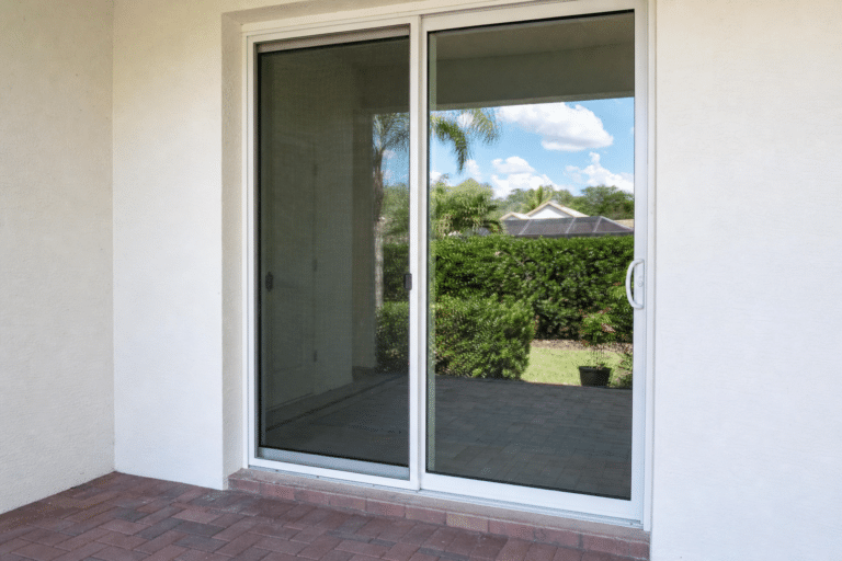 A closed glass sliding door opens onto a patio with brick flooring, showing a view of bushes, palm trees, and a partly cloudy sky outside.