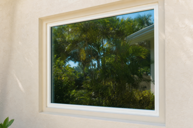 A clear window set in a beige stucco wall reflects green palm trees and blue sky from the outside environment.
