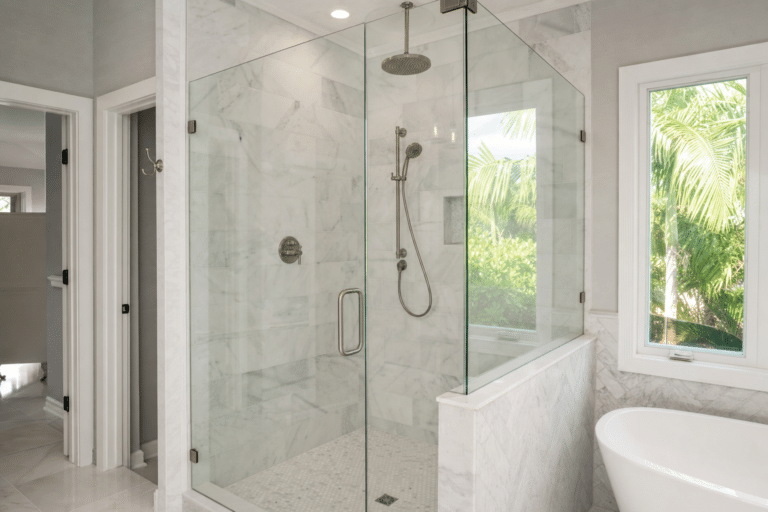 Modern bathroom with a glass-enclosed shower, marble walls and floors, a freestanding bathtub, and a window showing greenery outside.