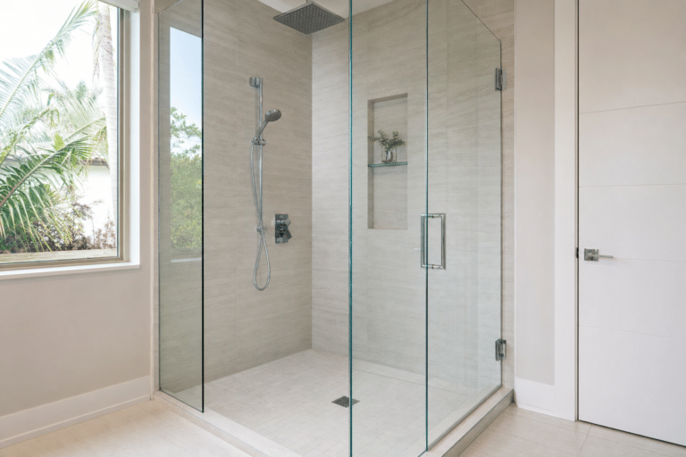 Modern glass-enclosed shower with a rainfall showerhead, handheld sprayer, built-in shelf, and beige tile walls. Large window and white door visible in the bathroom.