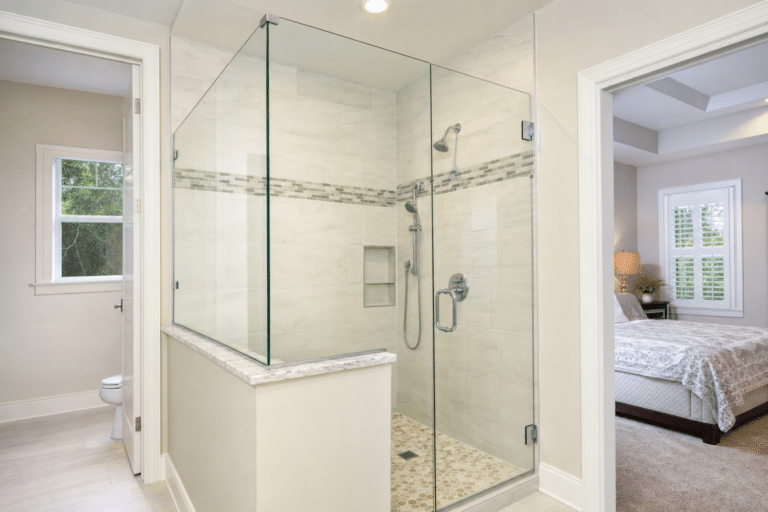 Modern bathroom with a glass-enclosed shower, white tile walls, built-in shelves, and chrome fixtures; toilet and bedroom visible in adjoining rooms.