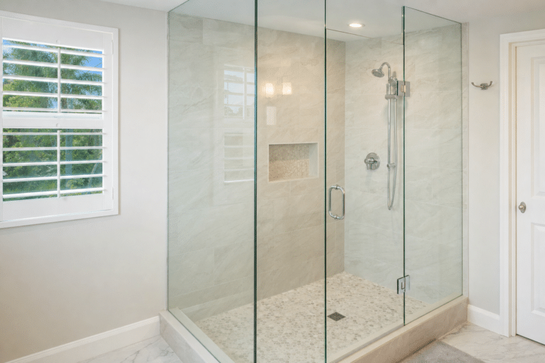 Modern bathroom with a large glass-enclosed walk-in shower, wall-mounted showerhead, built-in niche, and tiled walls and floor; sunlight coming through a window with white shutters.