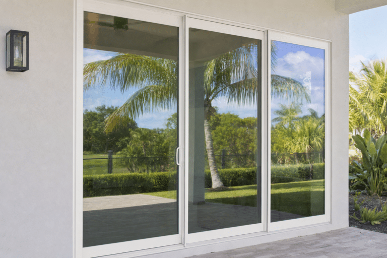Large glass sliding door reflects palm trees and green lawn outside, with a concrete patio and light-colored exterior wall visible.