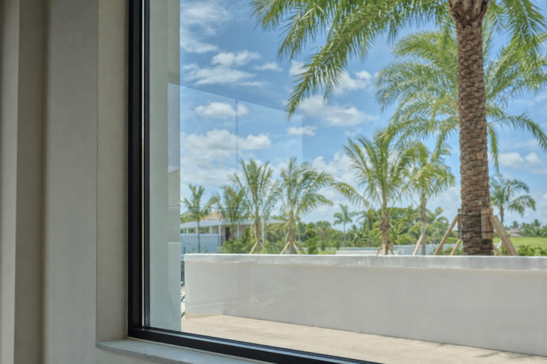 View through a window showing a patio with palm trees, a white wall, and a partly cloudy sky in the background.