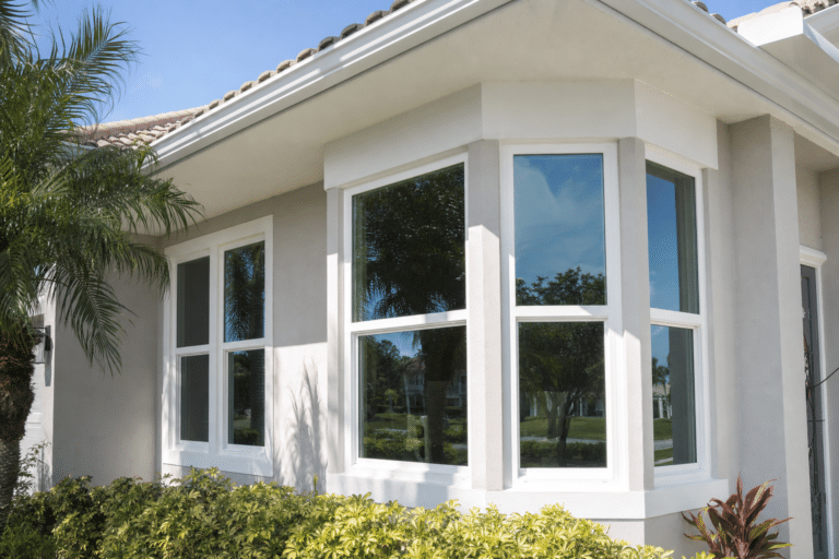 Exterior view of a house with three large white-framed windows, beige stucco walls, a tiled roof, and surrounding landscaping with green plants and a palm tree.