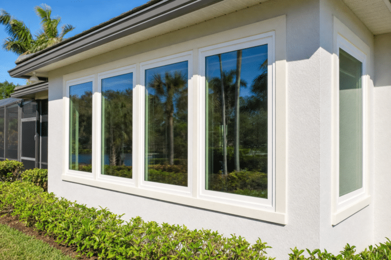 Four tall rectangular windows are set in a white stucco house exterior, reflecting palm trees and greenery on a sunny day.