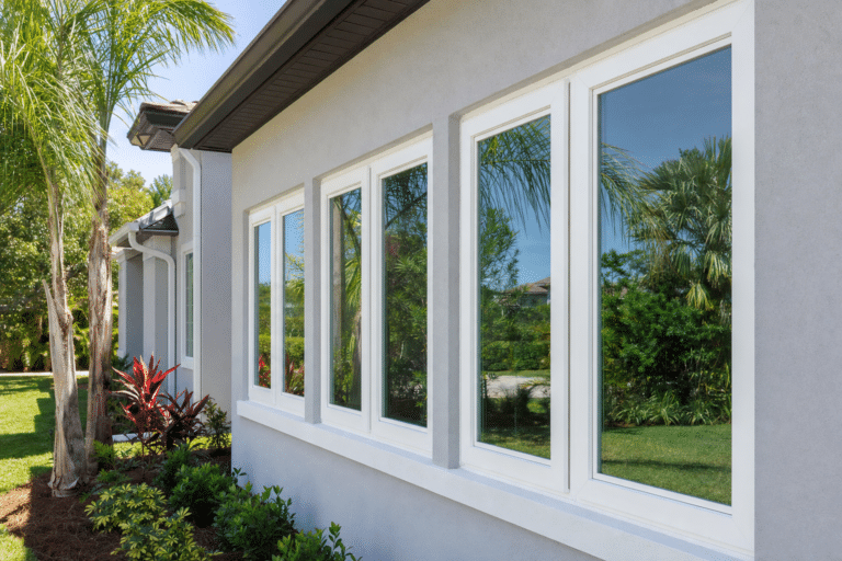 Row of white-framed windows on a gray house, reflecting palm trees and blue sky; landscaping with tropical plants is visible in the foreground.